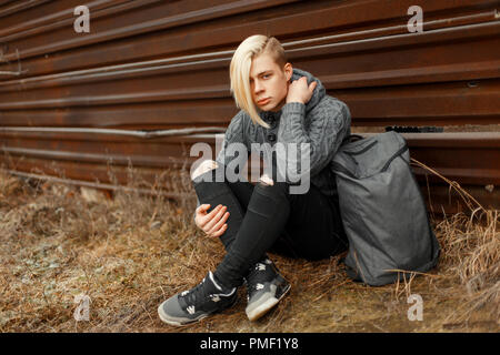 handsome young man with a haircut in a fashionable vintage knitted sweater with black jeans and sneakers with gray backpack sits near the metal wall Stock Photo