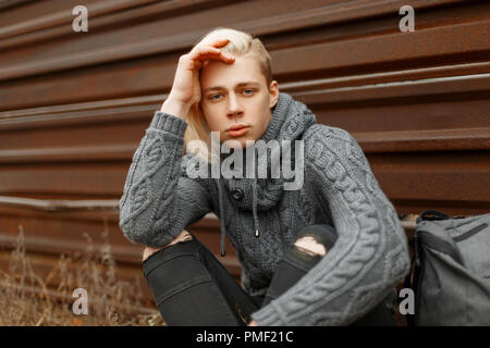 Stylish young man with a gray fashion sweater and a backpack sitting near a metal wall Stock Photo