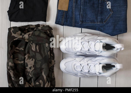 White sneakers, military cloth, a black T-shirt and blue jeans on a wooden floor. Fashionable stylish man's 2018 collections on a white wooden backgro Stock Photo