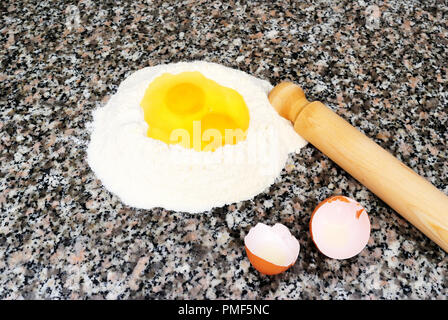 Flour fountain with two eggs in the center, near the broken shells and rolling pin, flat marble background, the ingredients to prepare desserts and do Stock Photo