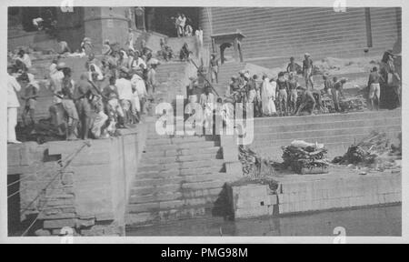 Black and white photograph on cardstock, with an image, at a slight angle, of a 'shmashana' or 'cremation' ghat, with several funeral pyres burning at the base of stone steps leading down to the Ganges river, and with a number of men, some arranging sticks for more funeral pyres, others sitting and standing in the background (including two men wearing white suits at far left) likely collected as a tourist souvenir during a trip to India, 1910. () Stock Photo