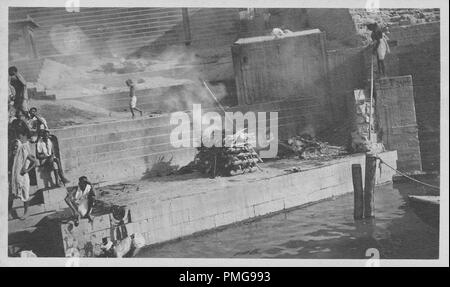 Black and white photograph on cardstock, with an image, shot from a high angle, of a 'shmashana' or cremation ghat, with a funeral pyre burning at the base of stone steps leading down to the Ganges river, several men, sitting and standing on the stairs at left, a young boy walking on a higher level behind the pyre, and a man at right holding a long stick, likely collected as a tourist souvenir during a trip to India, 1910. () Stock Photo