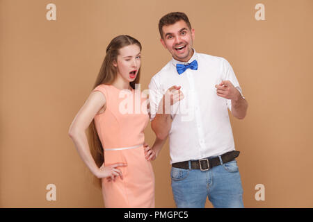 Portrait of handsome young man and beautiful woman in pink dress looking at camera with funny face, pointing and winking with amazed face. indoor stud Stock Photo