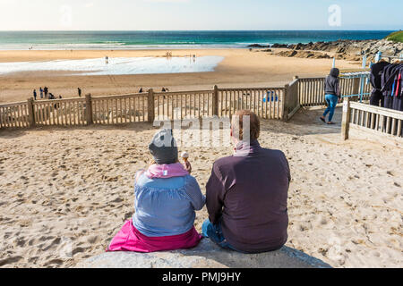 Holidaymakers on a staycation holiday enjoying an ice cream as they sit gazing out over Fistral Beach in Newquay in Cornwall. Stock Photo