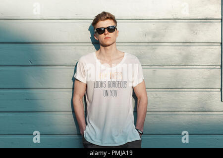 Handsome young stylish model man in sunglasses with a stylish white t-shirt stands near a wooden blue wall Stock Photo