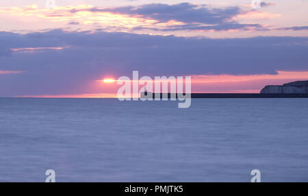 The harbour wall and Newhaven lighthouse is jutting out in the middle of the image behingd are chalk cliffs and a pink and blue sunset sky is behind a Stock Photo