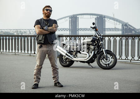 Redhead biker with beard in leather jacket standing with crossed arms near motorbike on bridge Stock Photo