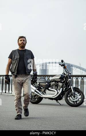 Redhead biker with beard in leather jacket standing walking on bridge near motorbike Stock Photo