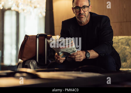 Portrait of confident mature businessman sitting in hotel foyer with his luggage on the side. CEO waiting in hotel lobby. Stock Photo