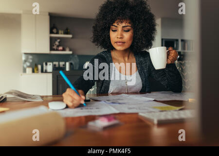Female architect making a project plan with cup of coffee in hand. Interior designer correcting a drawing while sitting at home office. Stock Photo