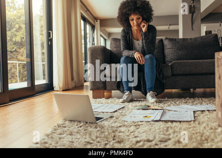Woman sitting on sofa looking at the papers lying on floor with a laptop. Woman working on a project from home. Stock Photo