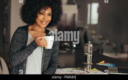 Happy woman taking coffee break while working at home office. African female sitting at her desk with cup of coffee looking away smiling. Stock Photo
