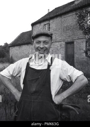 1950s, historical, portrait of a male estate maintenance man or handyman standing outside a farm property, wearing his work clothes, collared shirt, dunagarees and flat cap, England, UK. Stock Photo