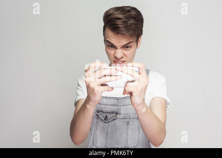 Angry crazy gamer teenager wearing in demin overalls and white T-shirt standing, using smartphone and playing mobile game with upset face. Indoor, iso Stock Photo