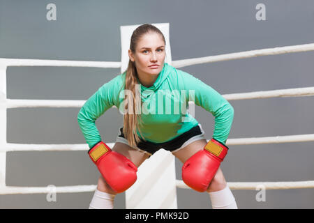 Angry boxing athletic young woman in green long sleeve poising with red boxing gloves and looking at camera with serious face. indoor studio shot on g Stock Photo