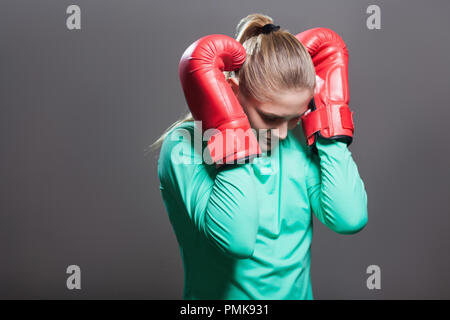 Sad young athlete woman with collected hair standing in position, holding head down and hand in boxing red gloves near the ears after knockout. Indoor Stock Photo