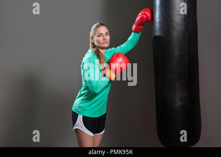 Portrait of serious young athlete woman with collected brunette hair standing and pushing punching bag in boxing red gloves and looking at camera. Ind Stock Photo