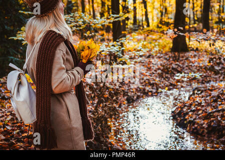 Rear view of female holding bouquet of yellow autumn maple leaves in her gloved hands. Ground covered with orange leaves lightened by warm evening backlit sun light reflected in a pond Stock Photo