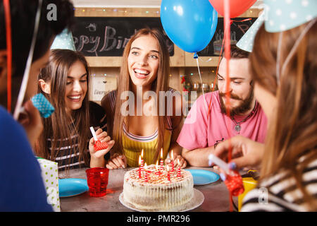 Five young adults celebrating a birthday party as they sitting around a table with cake and party hats Stock Photo