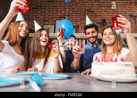 Five diverse group of friends at party sitting at the table in front of large birthday cake while holding drinks Stock Photo