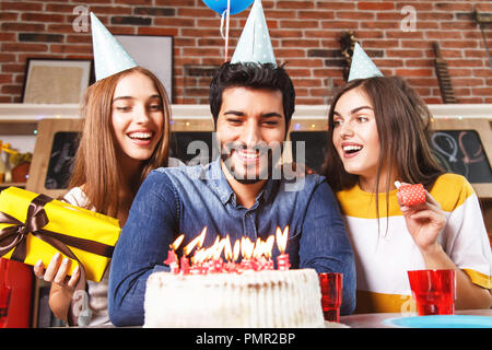 Happy bearded man ready to blow out candles on the white cake at birthday party with his girlfriends Stock Photo
