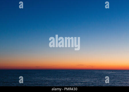 The glow of sunset after the sun has disappeared behind the horizon with deep blue sea in the foreground and a big empty blue sky Stock Photo