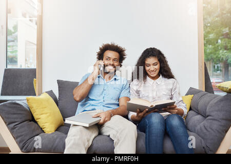 Portrait of two young good-looking dark-skinned students in casual clothes sitting together on sofa in modern library, reading books, preparing for exams, talking o phone. Stock Photo