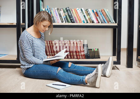 Young cheerful light-haired student girl with short hair in striped shirt and jeans sitting on floor in library, reading book, spending productive time after study, getting ready for exams. Stock Photo