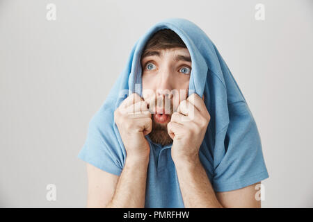 Funny and emotive caucasian guy hiding in shirt, staring through collar up as if seeing something terrifying and shocking, standing over gray background. Man went on kitchen and saw flying cockroach Stock Photo
