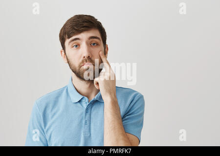 You have not impressed me. Studio shot of ordinary young bearded guy holding index finger on face, showing fake teardrop, standing with gloomy expression, imitating that he is sad over gray background Stock Photo