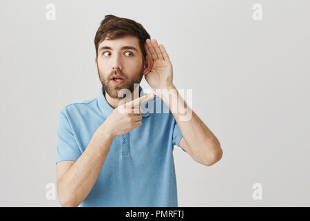 Are you talking about that person. Portrait of excited and shocked caucasian man holding hand near ear, pointing right with amazed expression, gossiping or spreading rumor about person who stands near Stock Photo