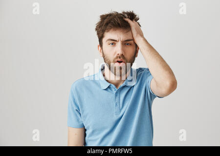 Funny mature bearded man with messy hair and beard, rubbing head and being tired or having hangover, standing over gray background. Confused and sleepy guy after celebrating b-day with fellows Stock Photo