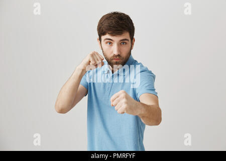 You have no chance to bit me. Studio shot of serious confident young bearded man standing in street fighting pose, raising fists and ready to battle over gray background. Hit back if you can Stock Photo