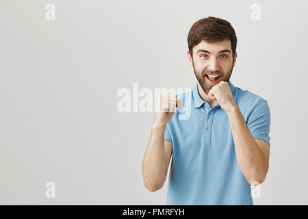 Happy joyful caucasian guy smiling while standing in defense pose with lifted fists that cover face from punch, being ready to fight over gray background. Bully enjoys get into fights Stock Photo