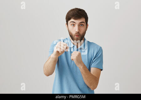 Punch from right and you are in knock out. Studio shot of funny emotive caucasian male with beard raising fists and pulling hand at camera as if hitting it, standing over gray wall in playful mood Stock Photo