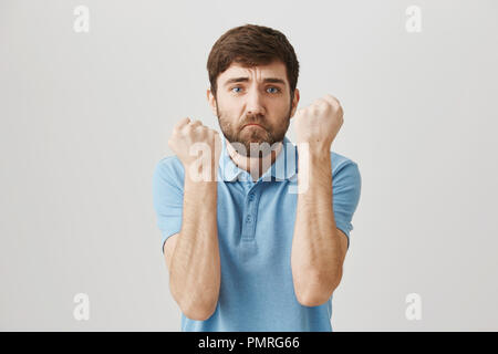 Mommy said I am strong. Studio shot of silly european guy being afraid while in middle of fight, raising fists, trying to protect himself from being bitten, standing over gray background. Stock Photo
