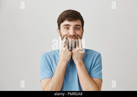 Gloomy guy with sad smile. Portrait of upset tired young bearded man stretching mouth with index fingers, looking bothered and exhausted while standing over gray background. Stock Photo