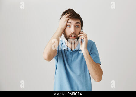 Man totally forgot about his meeting. Portrait of confused and shocked man holding hand on head and talking on smartphone, being concerned while standing against gray background. Stock Photo
