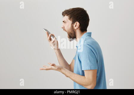 Stupid phone. Studio shot of angry crazy bearded guy shouting at smartphone while talking on speaker or being mad gadget is not working, standing over gray background in profile and gesturing Stock Photo