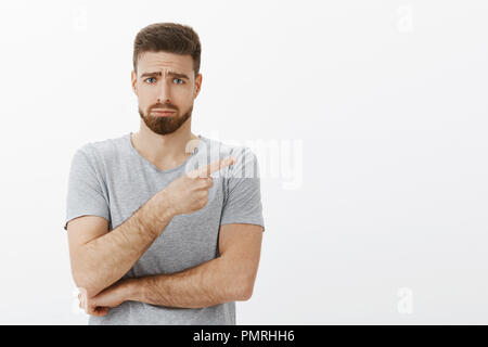 Sad and cute charming bearded male model in grey t-shirt frowning making gloomy face with frowned eyebrows, sulking pointing right expressing regret and envy standing unhappy over white background Stock Photo