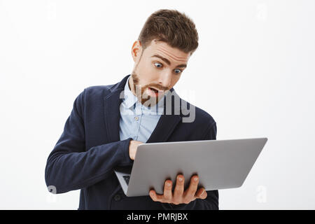 Man going crazy, being in rush working on project. Anxious troubled good-looking male with beard in suit holding laptop looking at computer screen, posing concerned and focused against grey wall Stock Photo