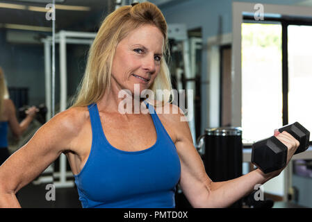 Female middle age fitness model shows muscular and lean arm strength while performing a dumbbell curl in gym. Stock Photo