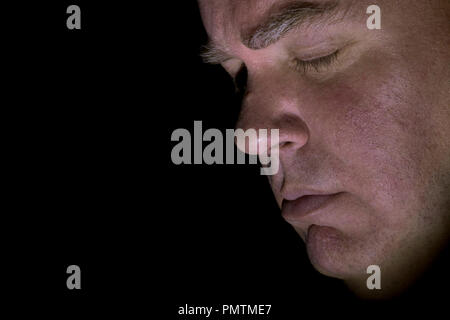 Model released closeup image of middle aged Caucasian man's face with eyes closed, looking sad and solemn, side view with black background. Stock Photo