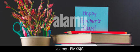Concept of Teacher's Day. Objects on a chalkboard background. Books, green apple, plaque: Happy Teacher's Day, pencils and pens in a glass, sprig with Stock Photo