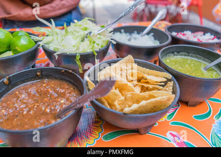 Isolated, close-up of red salsa, green salsa, limes, tortilla chips, shredded cabbage, onions, radishes, in black bowls with a colorful tablecloth Stock Photo