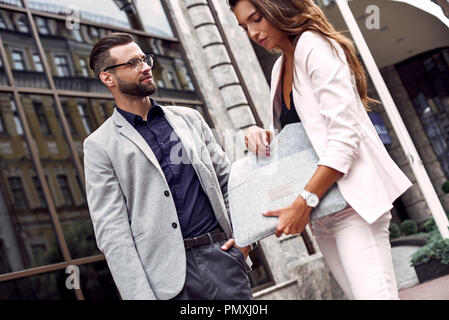 Waiting for colleague. Two young business people standing outside on the city street man looking curious at woman taking documents from her bag close-up Stock Photo
