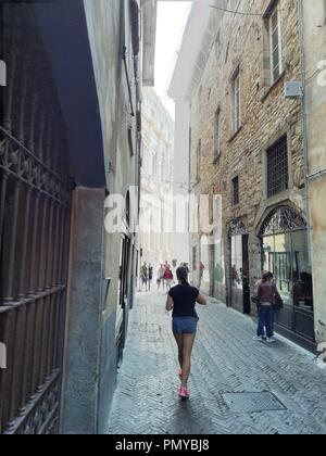 Italy, the historic and charming city of Bergamo. A young woman walks towards the old city square, down a shaded narrow street with tall buildings. Stock Photo