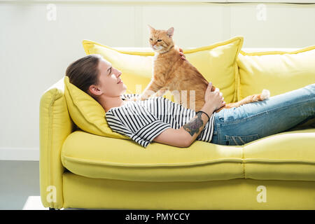 beautiful girl lying on yellow sofa and palming cute red cat in living room Stock Photo