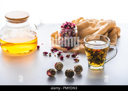 dried rose buds in jar, cup of chinese flowering tea with tea balls on white tabletop Stock Photo