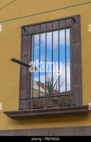 Looking through a stone window frame with bars, a stone window sill, with a cloudy blue sky and a potted plant, surrounded by a yellow painted wall Stock Photo